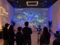 Concept render: visitors in The Rectangle watching a robot guide present the projected Agent City map on the wall